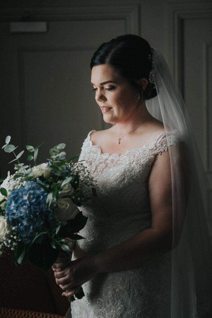 a feminine portrait of bride standing by the window and softly looking at her flowers