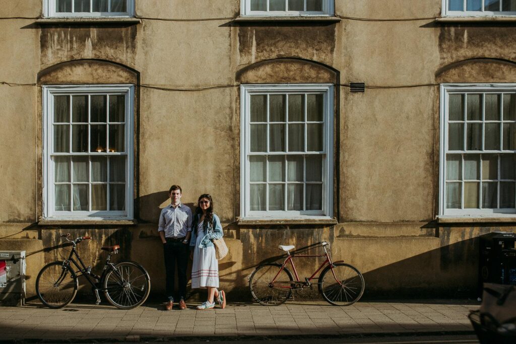 a couple is leaning against the building wall and looking directly on the camera.The setting sun is casting long shadows on the street of oxford