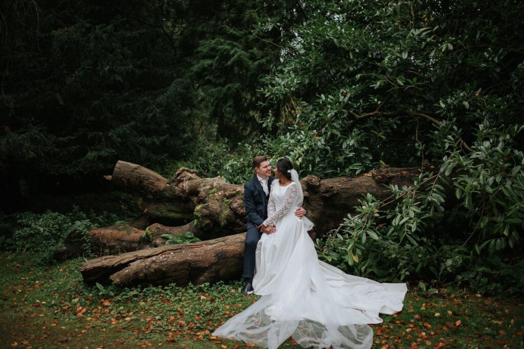 bride and groom are sitting on the large fallen tree trunk. They are surrounded by the deep green bushes