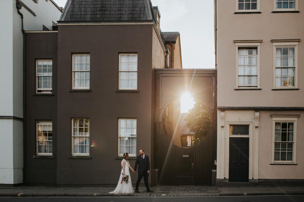 bride and groom walking on the st. Aldates street in oxford. The sun rays are penetrating gaps in the road gate which create a lovely flare on the image