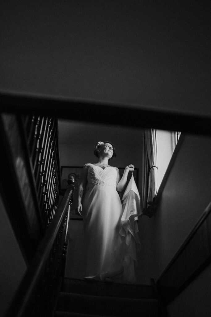 black and white image of bride ready to leave for the church standing on the top of the staircase.