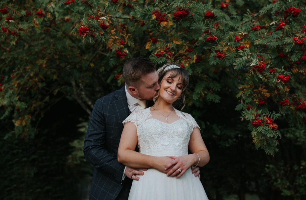 groom is gently kissing brides cheek. there is a stunning rowan ash tree in the backdrop