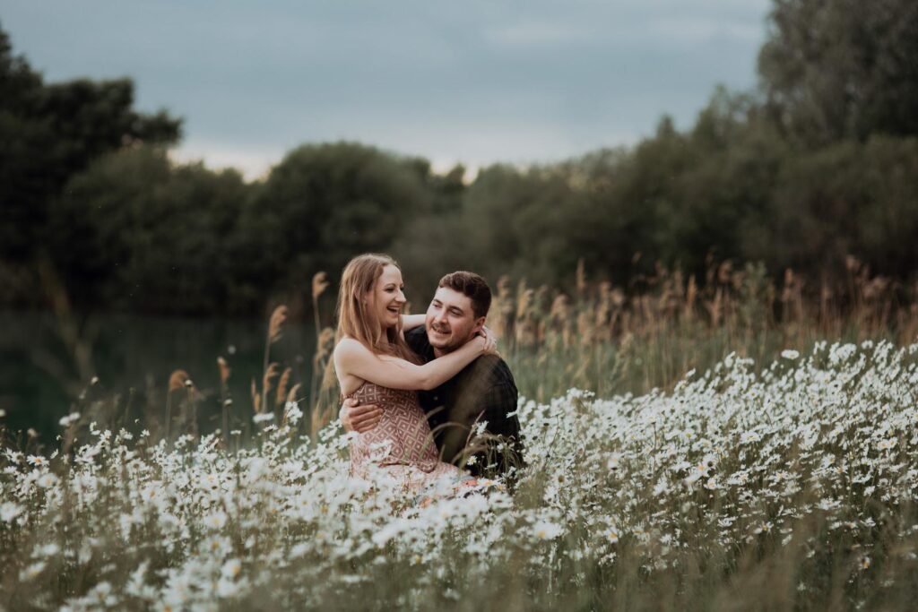 a man and woman standing on the meadow surrounded by the booming daisies. He is picking her up