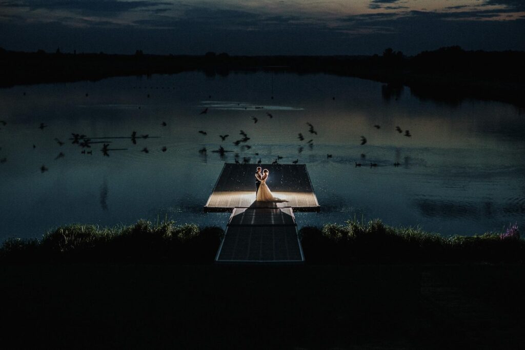 atmospheric night image of bride and groom standing on the footbridge by the lake. in the background a flock of goose is landing on water