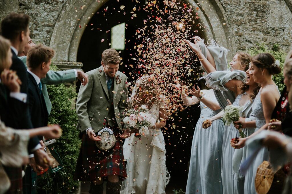 bride and groom who is wearing Scottish kilt leaving the church. wedding guests joyfully throwing confetti made of dried rose petals