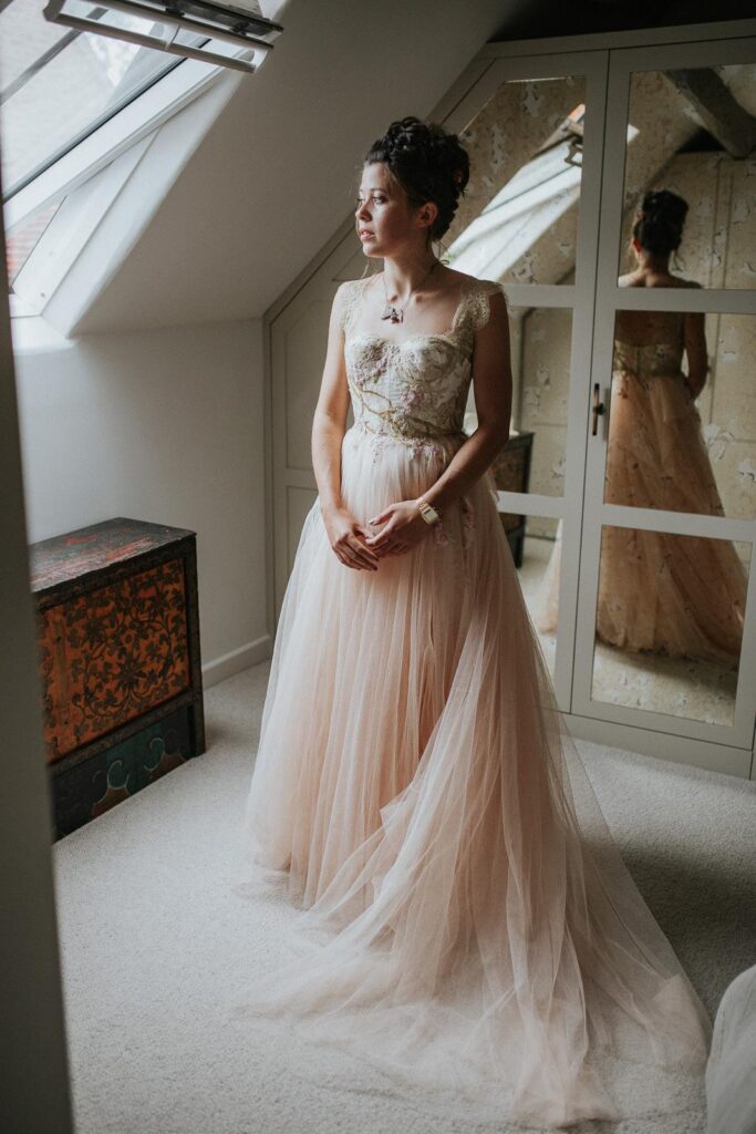 a bride wearing beautiful pink wedding dress is standing by the window of her bedroom